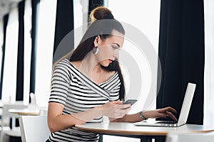 Charming Caucasian woman using mobile phone and laptop computer while sitting in modern coffee shop interior, young