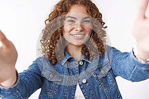 Charming charismatic happy redhead curly-haired girl freckles pimples extend arms forward grab camera as taking selfie