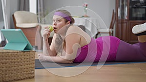 Charming confident obese Caucasian woman lying on exercise mat eating healthful vitamin apple. Side view portrait of