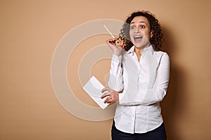 Charming curly woman in a white shirt stands on a beige background with a pen and a diary , looking up with opened mouth