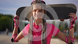 Charming cute girl holding skateboard behind head smiling and looking away in slow motion. Portrait of happy confident