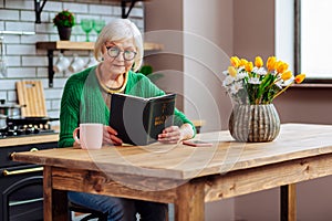 Charming dame with silver hair reading Bible at kitchen table