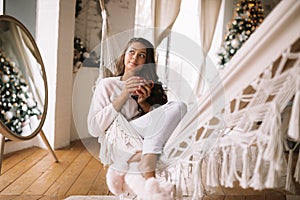 Charming dark-haired girl dressed in pants, sweater and warm slippers holds a red cup lying in a hammock in a cozy