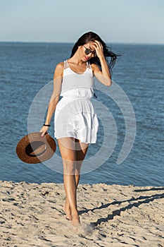 Charming dark-haired girl in white dress and sunglasses hold hat in hand and walk on the sandy beach near the sea on a