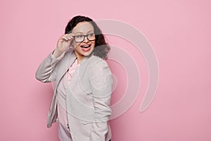 Charming dark-haired woman smiling, looking aside, wearing trendy glasses and elegant light gray suit, pink background