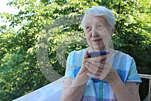 Charming elderly woman in rectangular black rimmed eyewear using voice recognition function on cell phone.