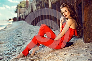 Charming elegant young woman in coral red one shoulder jumpsuit sitting on the beach at the old rusty piles