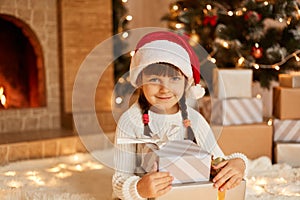 Charming female kid holding stack of present, little kid wearing white sweater and santa claus hat, sitting on floor near