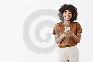 Charming and friendly African American teenage girl with curly hair in stylish outfit holding smartphone and smiling