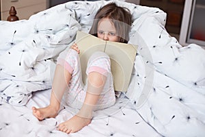 Charming girl reading her favorite book. Cute little child reading fairy tale and looks concentrated, lying in bed, holding book