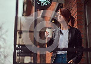 A charming girl wearing a leather jacket with a rucksack holding cup with takeaway coffee outside near the cafe.