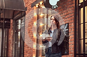 A charming girl wearing a leather jacket with a rucksack holding cup with takeaway coffee outside near the cafe.