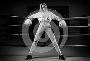 Charming girl in a white sweatshirt poses with huge white gloves against the background of a boxing ring. The concept of sports,