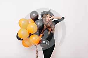 Charming graceful girl in witch hat holding helium balloons. Indoor shot of blithesome brunette lady in long dress