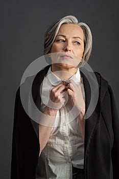 Charming gray-haired woman thoughtfully looks away adjusting collar of white shirt, throwing black jacket over her