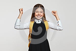 Charming happy girl with yellow backpack wearing school uniform, raise hands up in good mood winner