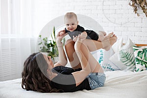 Charming happy little baby boy having fun with mom brunette woman on bed in the bright bedroom