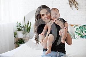 Charming happy little baby boy having fun with mom brunette woman on bed in the bright bedroom