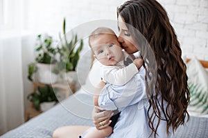 Charming happy little baby boy having fun with mom brunette woman on bed in the bright bedroom