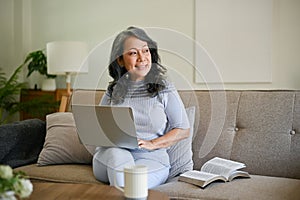 A charming and happy middle-aged Asian woman working on her tasks on her laptop on a sofa