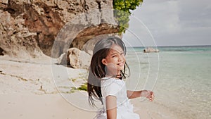 A charming and happy philippine teenage girl in a white summer dress is running along a tropical beach near the rocks