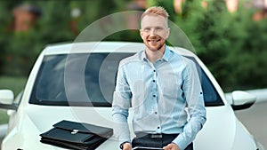 Charming happy positive young business man posing outdoor looking at camera sitting on car bonnet