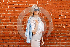 Charming happy young hipster woman with a beautiful smile in a white lace blouse in a denim jacket in black sunglasses