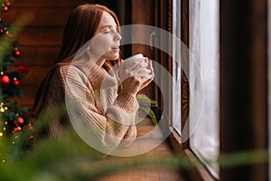 Charming happy young woman looking out of window and drinking with enjoying hot coffee at home