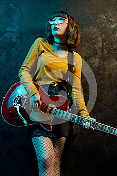 Charming hipster woman with curly hair with red guitar in neon lights. Rock musician is playing electrical guitar. 90s