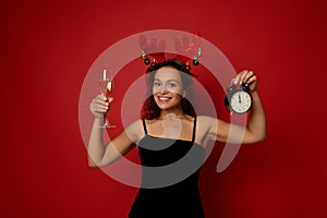 Charming Hispanic woman wearing antler hoop and black velvet dress holding an alarm clock and champagne flute with sparkling wine