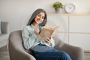 Charming Indian lady with cup of hot coffee and open book sitting in armchair at home