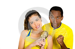 Charming interracial couple wearing yellow football shirts holding small trophy posing for camera, white studio