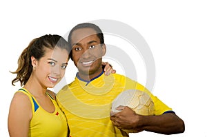 Charming interracial couple wearing yellow football shirts, hugging friendly while posing for camera holding ball, white