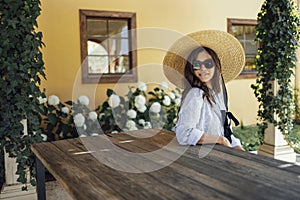 Charming Italian girl in casual dress and wide brimmed straw hat sitting on beanch at table on terrace photo