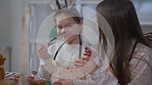 Charming joyful girl coloring nose with food paint sitting at table with sister. Portrait of cheerful cute Caucasian