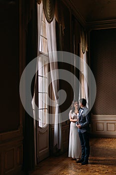 Charming just married couple lovingly embracing near large illuminated window in vintage victorian mansion