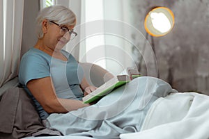 Charming lady lying in bed and engaging in book reading.