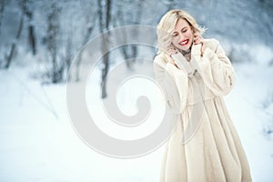 Charming laughing blond lady with perfect make up wearing luxurious white swing fur coat standing on winter snowy background