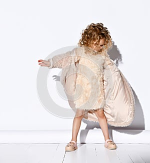 Charming little blond girl with curly hair in pink dress and sandals getting into fashionable fuzzy coat.