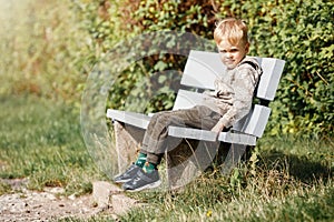 Charming little boy calmly sitting on the wooden bench in the city park