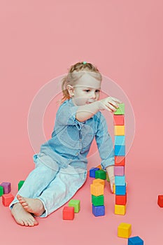 A charming little girl in denim clothes on a pink background builds a tower