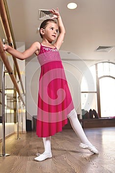 Charming little girl dreams of becoming a ballerina. The girl in the pink dress is dancing, holding on to the bar.Baby girl is