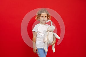 Charming little girl hold in hand favourite stuffed toy on empty red background, free copy space. Photo of nice child of