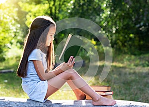 Charming little girl with long brown hair reads book outdoor sitting on tree in summer park or in a forest glade. booklover