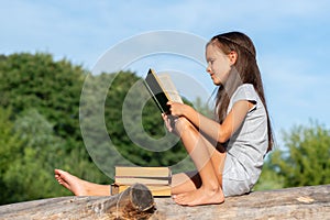 Charming little girl with long brown hair reads book outdoor sitting on tree in summer park or in a forest glade. Booklover