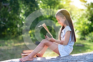 Charming little girl with long brown hair reads book outdoor sitting on tree in summer park or in a forest glade. Booklover