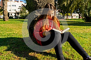 Charming little girl with long brown hair reads book outdoor sitting on tree in winter park or in a forest glade. Child