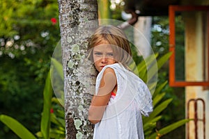 Charming little girl standing near the tree i tropical garden, wearing white dress. Sad face with pensive emotion