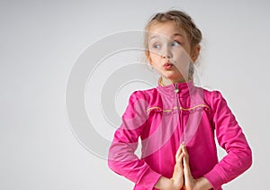 Charming little girl stands clasping hands, looking aside and funnily pursing her lips . Waist-up studio shot isolated