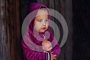 Charming little girl in a warm burgundy jacket, sitting on the street with a doll in her hands. Portrait of a pensive